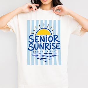 May include: Cream-colored t-shirt featuring a blue and yellow graphic. The design includes a yellow sun, blue waves, and the words "SENIOR SUNRISE CLASS OF 2027" in blue. The background has vertical light blue stripes.