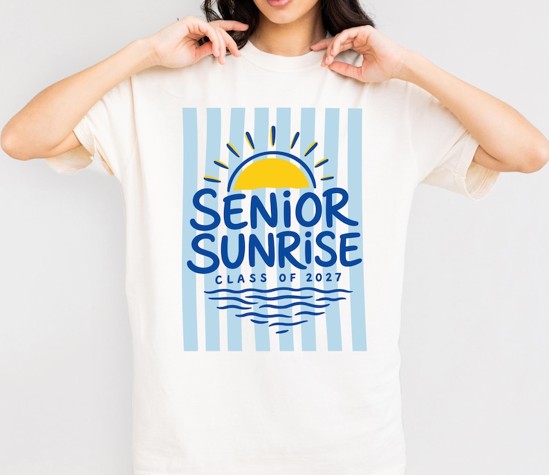 May include: Cream-colored t-shirt featuring a blue and yellow graphic. The design includes a yellow sun, blue waves, and the words "SENIOR SUNRISE CLASS OF 2027" in blue. The background has vertical light blue stripes.
