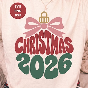 Könnte beinhalten: Ein cremefarbenes T-Shirt mit einer Grafik einer Weihnachtskugel mit einer rosa Schleife. Das Wort CHRISTMAS ist in Rot gedruckt, und das Jahr 2026 steht darunter in Grün. Ein roter Kreis oben links enthält den Text SVG PNG DXF.