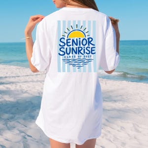 May include: White t-shirt with a graphic on the back featuring a yellow sun, blue waves, and the words "SENIOR SUNRISE CLASS OF 2027" in blue. The design is set against a backdrop of light blue vertical stripes.