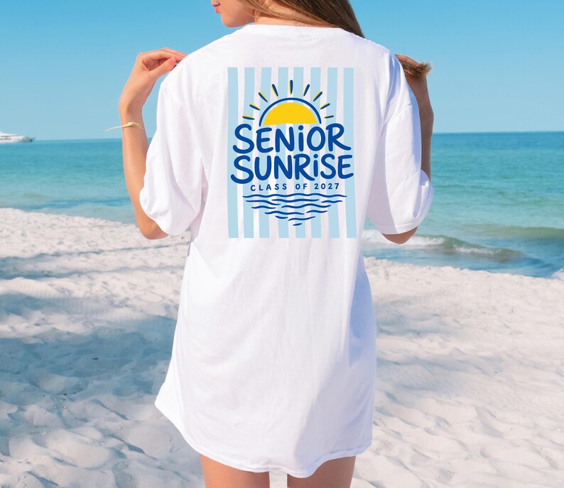 May include: White t-shirt with a graphic on the back featuring a yellow sun, blue waves, and the words "SENIOR SUNRISE CLASS OF 2027" in blue. The design is set against a backdrop of light blue vertical stripes.