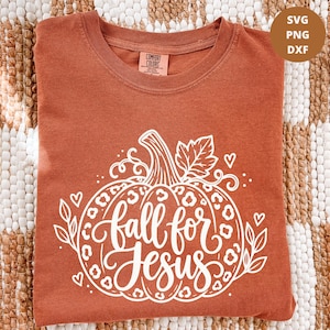 May include: A rust-colored t-shirt featuring a white graphic of a pumpkin with a leopard print pattern. The text "fall for Jesus" is written in a cursive script across the center of the pumpkin, surrounded by small hearts and leaf accents.