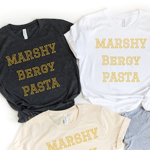May include: A collection of t-shirts in various colors, including white, black, gray, and beige. Each shirt features the text "MARSHY BERGY PASTA" in a gold, collegiate-style font. The shirts are short-sleeved and appear to be made of a soft material.