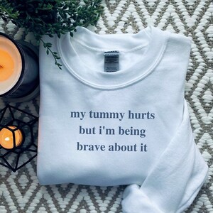 Könnte beinhalten: Weißer Crewneck-Pullover mit dem Aufdruck "my tummy hurts but i'm being brave about it" in Blau.