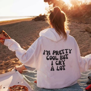 Puede incluir: Una sudadera blanca con un gráfico negro que dice "I'm pretty cool but I cry a lot" en la espalda. La sudadera la lleva puesta una persona sentada en una playa con una caja de pizza y una rebanada de pizza.