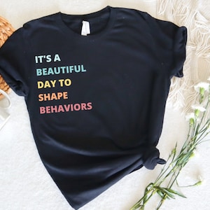 以下が含まれることがあります： 虹色の文字で「It's a beautiful day to shape behaviors」と書かれた黒いTシャツです。