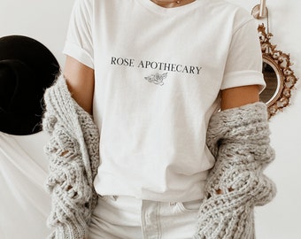 Puede incluir: Camiseta blanca con el texto "ROSE APOTHECARY" y un pequeño diseño floral.