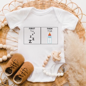 Peut inclure: Combinaison blanche pour bébé avec le texte "FIRST Nap zzzzzz THEN Milk" et un graphique d'un biberon avec une tétine rouge.