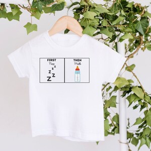 May include: White baby onesie with a graphic of two boxes. The first box says "First Nap" with a series of Z's. The second box says "Then Milk" with a drawing of a baby bottle.