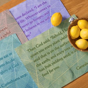 May include: Four placemats with a leaf pattern and inspirational bible verses. The placemats are in shades of green, blue, and purple. The verses are from the books of Corinthians and Genesis.