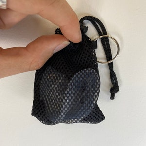 Puede incluir: Una bolsa de cordón de malla negra con un anillo plateado y un cordón negro. La bolsa está llena de pequeños objetos negros redondos.