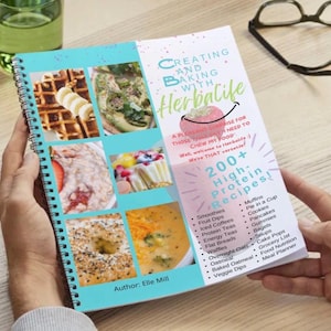 Op de afbeelding: Een spiraalgebonden kookboek getiteld "Creating and Baking with Herbalife" wordt opengehouden en toont kleurrijke foto's van voedsel en een lijst met recepten. De omslag toont het Herbalife-logo en de naam van de auteur, Elle Mill. Het boek belooft meer dan 200 eiwitrijke recepten.