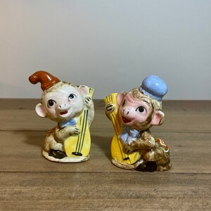 以下が含まれることがあります： 音楽の楽器を演奏しているセラミック製の猿の置物2体。左側の猿は茶色の帽子をかぶり、右側の猿は青い帽子をかぶっています。両方の猿は黄色の楽器を持っています。