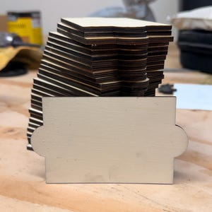 May include: A stack of laser-cut wooden coasters with a dark edge and a light-colored top. A single coaster is in the foreground, painted in a light cream color. The coasters are stacked on a wooden surface.