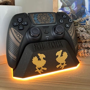 K&ouml;nnte beinhalten: Ein schwarzer PlayStation 5 DualSense-Controller mit einem goldenen und blauen geometrischen Design auf dem Touchpad. Der Controller sitzt auf einem schwarzen Ladest&auml;nder mit einem goldenen Final Fantasy-Logo und zwei goldenen Chocobo-Figuren. Der St&auml;nder hat ein leuchtend orangefarbenes Licht um die Basis.