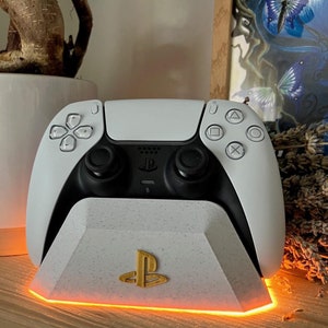 Peut inclure: Une manette de jeu PlayStation 5 blanche est posée sur un socle de charge blanc avec un logo PlayStation doré. Le socle est doté d'un éclairage orange lumineux autour de sa base.