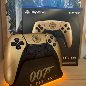 Op de afbeelding: Een goudkleurige PlayStation 5-controller met zwarte knoppen en accenten, weergegeven op een zwarte standaard met de tekst "007 FIRST LIGHT". De controller staat voor een doos met de PlayStation- en Sony-logo's.