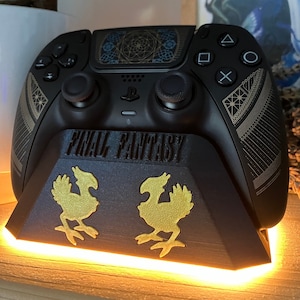 May include: A black PlayStation 5 controller with a gold and black design featuring a geometric pattern and the words "FINAL FANTASY". The controller is on a black stand with two gold Chocobo figures.