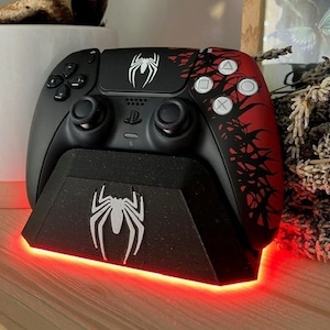 Op de afbeelding: Een zwarte PlayStation 5 DualSense-controller met een rood en zwart spinnenwebontwerp. De controller staat op een zwarte standaard met een wit spinnenlogo. De standaard heeft een rode LED-lichtstrip aan de onderkant.
