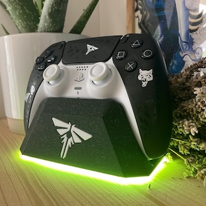 May include: A white and black video game controller with a black and white firefly design on the top and a wolf design on the back. The controller is sitting on a black charging stand with a green light.