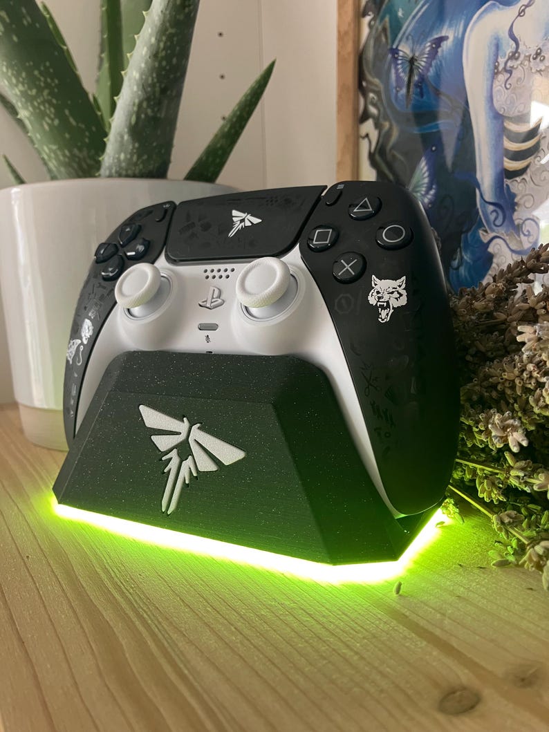 May include: A white and black video game controller with a black and white firefly design on the top and a wolf design on the back. The controller is sitting on a black charging stand with a green light.