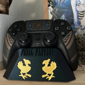 K&ouml;nnte beinhalten: Ein schwarzer PlayStation 5 DualSense-Controller mit einem goldenen und schwarzen geometrischen Design auf dem Touchpad. Der Controller steht auf einem schwarzen St&auml;nder mit den Worten "FINAL FANTASY" und zwei goldenen Chocobo-Figuren.