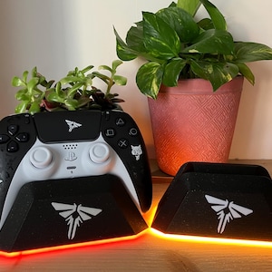 Puede incluir: Un mando de videojuego blanco y negro con una base de carga blanca. La base tiene una luz naranja brillante y un logotipo de luciérnaga blanco. El mando tiene un logotipo de luciérnaga blanco en la parte delantera y un logotipo de lobo blanco en la parte trasera.