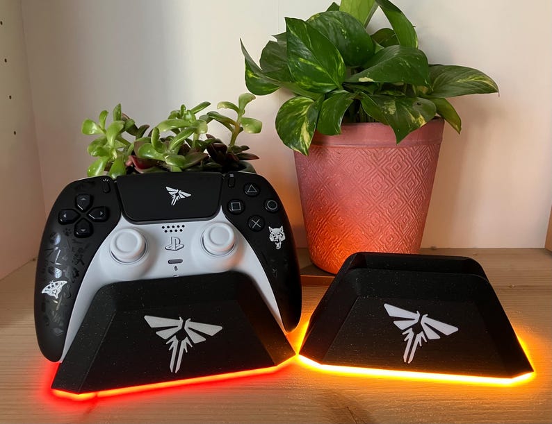 May include: A black and white video game controller with a white charging stand. The stand has a glowing orange light and a white firefly logo. The controller has a white firefly logo on the front and a white wolf logo on the back.