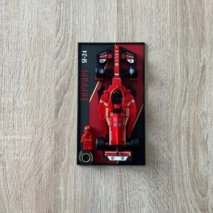 Peut inclure: Un modèle de voiture de course Lego Ferrari SF-24 rouge avec un cadre noir et une figurine Lego de pilote de course. Le modèle est présenté sur un fond noir.