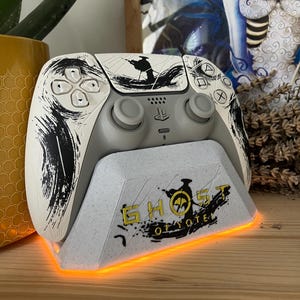 Peut inclure: Une manette PlayStation 5 blanche et grise avec un design personnalisé avec des coups de pinceau noirs et une silhouette. La manette repose sur un support assorti avec le texte "GHOST OF TSUSHIMA" en jaune. Le support a une base orange lumineuse.