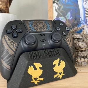 K&ouml;nnte beinhalten: Ein schwarzer PlayStation 5 DualSense-Controller mit einem gold-schwarzen Design, das ein geometrisches Muster auf dem Touchpad aufweist. Der Controller befindet sich auf einem schwarzen St&auml;nder mit dem Aufdruck "FINAL FANTASY" auf der Vorderseite. Der St&auml;nder hat zwei goldene Chocobo-Figuren auf der Vorderseite.