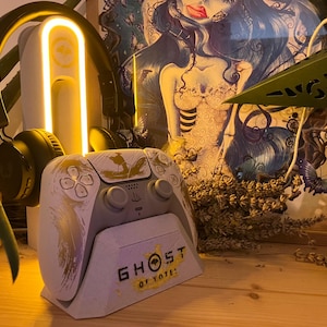Peut inclure: Une manette de jeu vidéo blanche et grise avec des détails dorés et le texte "GHOST OF YOTE!". La manette est sur un support. Un casque noir et une lumière au néon sont en arrière-plan.