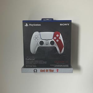 Puede incluir: Un mando inalámbrico DualSense de PlayStation blanco y rojo en su caja original. La caja presenta el logotipo de PlayStation, el logotipo de Sony y las palabras "GOD OF WAR 20TH ANNIVERSARY". El mando está diseñado para PS5, PC y Mac.