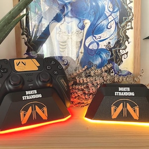 Op de afbeelding: Zwarte PlayStation 5 controller en oplaadstation set. De controller en het oplaadstation hebben oranje accenten en de woorden "DEATH STRANDING" erop gedrukt. Het oplaadstation heeft een rood gloeiend licht.