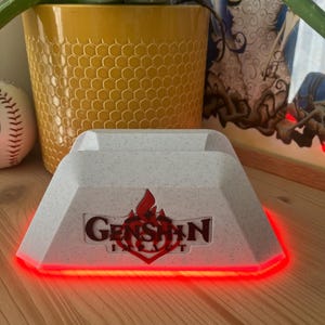 Peut inclure: Un support de chargement de console de jeu blanc et gris avec le mot "Genshin" en lettres rouges et un logo de flamme rouge. Le support a une lumière LED rouge autour de la base. Un pot jaune à motif en nid d'abeille avec une plante est en arrière-plan.