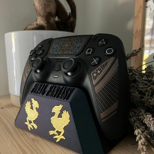 K&ouml;nnte beinhalten: Ein schwarzer PlayStation 5 DualSense-Controller mit einem goldenen Final Fantasy VII Remake-Design steht auf einem schwarzen St&auml;nder mit goldenen Final Fantasy VII Remake-Logos. Der St&auml;nder steht auf einer Holzoberfl&auml;che.