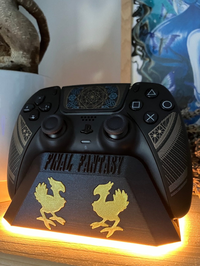 K&ouml;nnte beinhalten: Ein schwarzer PlayStation 5-Controller mit einem gold-schwarzen Design, das ein geometrisches Muster und die Worte "FINAL FANTASY" zeigt. Der Controller steht auf einem schwarzen St&auml;nder mit zwei goldenen Chocobo-Figuren.