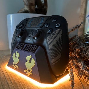 K&ouml;nnte beinhalten: Ein schwarzer PlayStation 5 DualSense-Controller mit goldenem und blauem Design steht auf einem schwarzen Ladest&auml;nder mit goldenem Final Fantasy-Logo und zwei goldenen Chocobo-Figuren. Der St&auml;nder hat ein wei&szlig;es LED-Licht um die Basis.