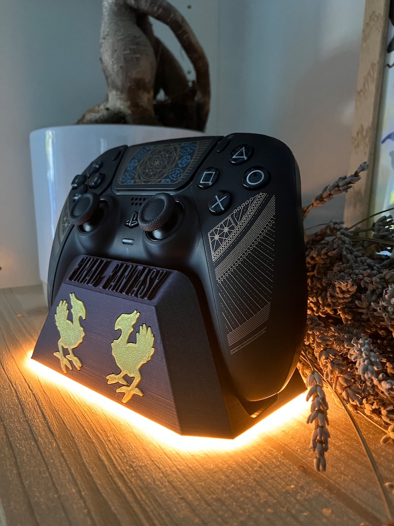 K&ouml;nnte beinhalten: Ein schwarzer PlayStation 5 DualSense-Controller mit goldenem und blauem Design steht auf einem schwarzen Ladest&auml;nder mit goldenem Final Fantasy-Logo und zwei goldenen Chocobo-Figuren. Der St&auml;nder hat ein wei&szlig;es LED-Licht um die Basis.