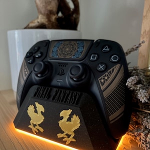 K&ouml;nnte beinhalten: Ein schwarzer PlayStation 5-Controller mit goldenem und schwarzem Design steht auf einem schwarzen St&auml;nder mit goldenem Final Fantasy-Logo und zwei goldenen Chocobo-Figuren. Der St&auml;nder hat ein leuchtend orangefarbenes Licht um die Basis.