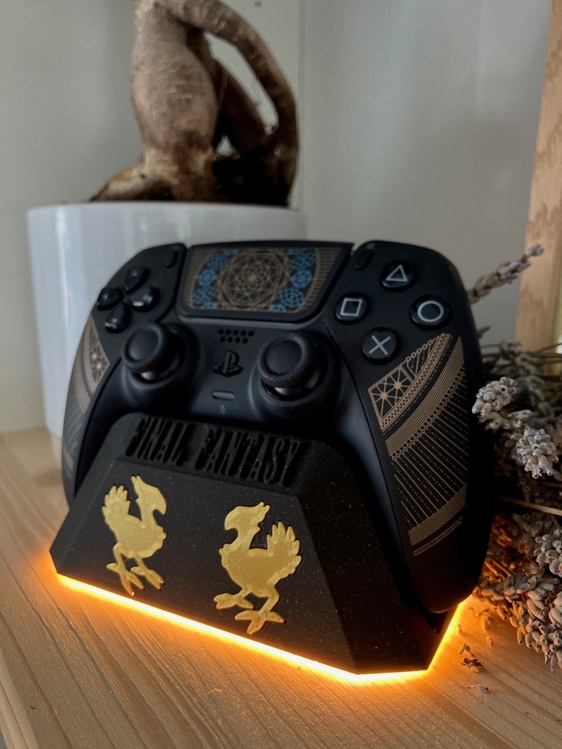 K&ouml;nnte beinhalten: Ein schwarzer PlayStation 5-Controller mit goldenem und schwarzem Design steht auf einem schwarzen St&auml;nder mit goldenem Final Fantasy-Logo und zwei goldenen Chocobo-Figuren. Der St&auml;nder hat ein leuchtend orangefarbenes Licht um die Basis.