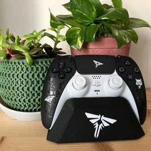 May include: A black and white PlayStation 5 DualSense controller with a white Firefly logo on the top and a white wolf logo on the right side. The controller is sitting on a black stand with a white Firefly logo on the front. The stand is on a wooden surface.