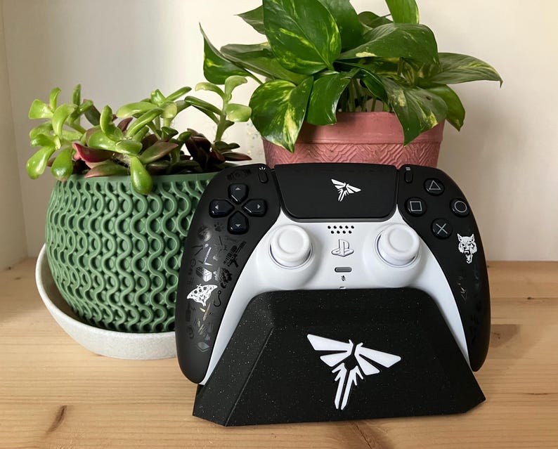 May include: A black and white PlayStation 5 DualSense controller with a white Firefly logo on the top and a white wolf logo on the right side. The controller is sitting on a black stand with a white Firefly logo on the front. The stand is on a wooden surface.