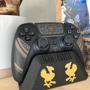 K&ouml;nnte beinhalten: Ein schwarzer PlayStation 5 DualSense-Controller mit einem geometrischen Design in Gold und Schwarz. Der Controller steht auf einem schwarzen St&auml;nder mit den Worten "FINAL FANTASY" und zwei goldenen Chocobo-Figuren.