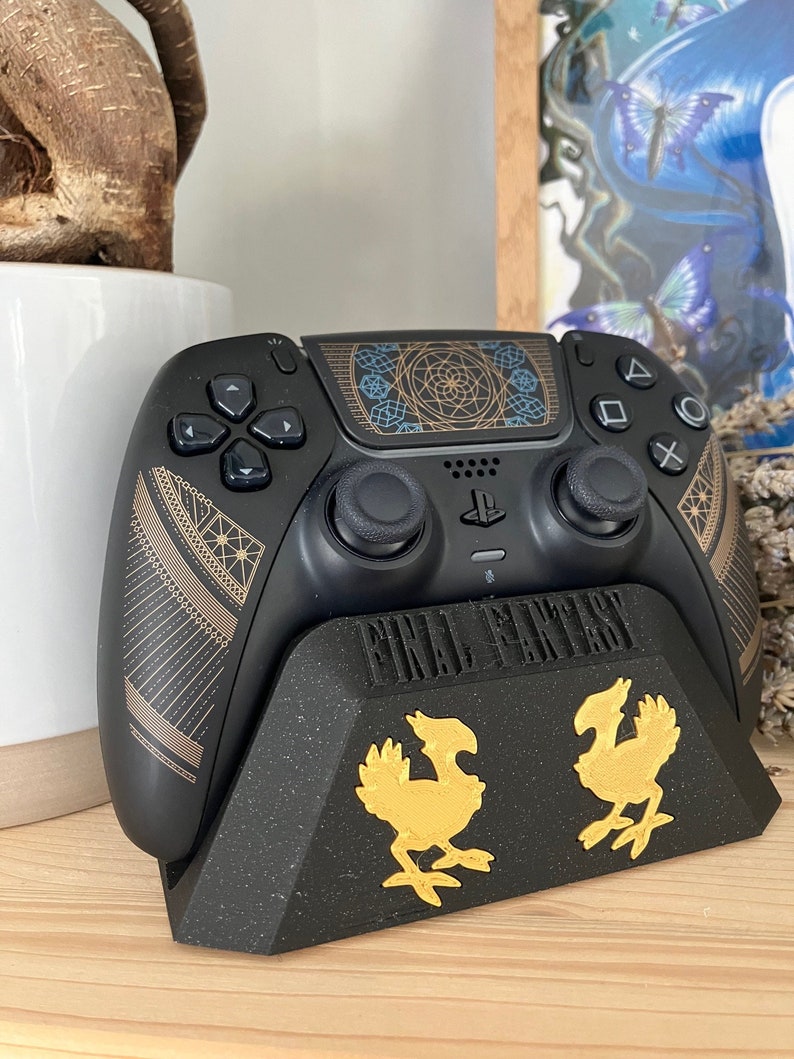 K&ouml;nnte beinhalten: Ein schwarzer PlayStation 5 DualSense-Controller mit einem geometrischen Design in Gold und Schwarz. Der Controller steht auf einem schwarzen St&auml;nder mit den Worten "FINAL FANTASY" und zwei goldenen Chocobo-Figuren.
