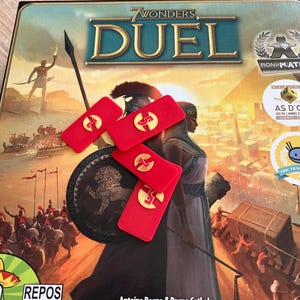 Peut inclure: Le jeu de société "7 Wonders Duel" est présenté, avec une illustration colorée de civilisations antiques. Des pions rouges avec des détails dorés sont disposés sur le plateau. La boîte du jeu est visible, avec le titre et les noms des créateurs.
