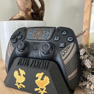 K&ouml;nnte beinhalten: Ein schwarzer PlayStation 5-Controller mit einem gold-schwarzen Final Fantasy-Design wird auf einem schwarzen St&auml;nder mit einem goldenen Final Fantasy-Logo und zwei goldenen Chocobo-Figuren pr&auml;sentiert.