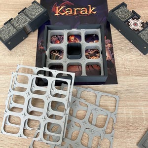 May include: A board game titled "Karak" with various components. The game includes a box, game board, gray plastic inserts, and gray plastic tiles. The game board features a dark purple background with the game title in gold lettering.