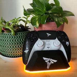 May include: A white and black video game controller with a black charging stand. The stand has a glowing orange light around the base and a white firefly logo on the front. The controller has a white firefly logo on the top and a white wolf logo on the side.
