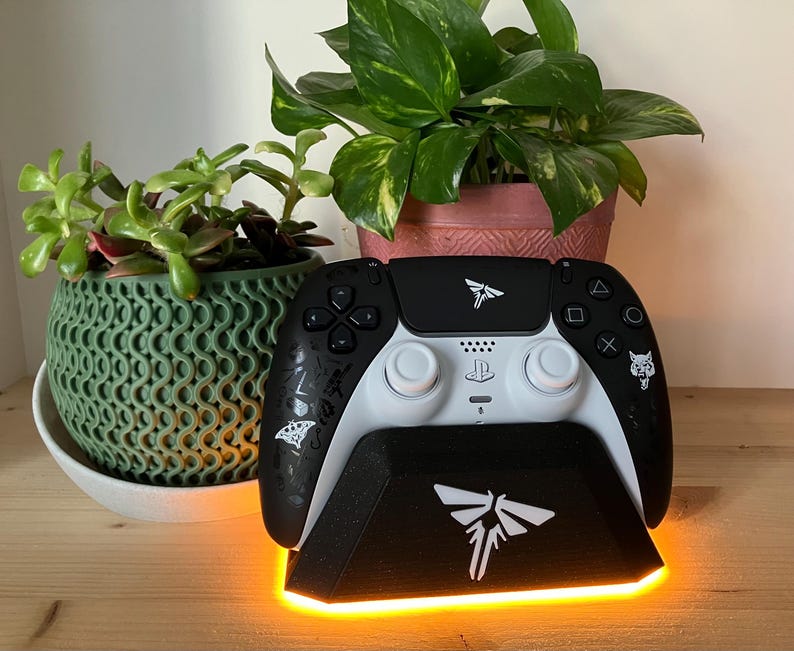 May include: A white and black video game controller with a black charging stand. The stand has a glowing orange light around the base and a white firefly logo on the front. The controller has a white firefly logo on the top and a white wolf logo on the side.